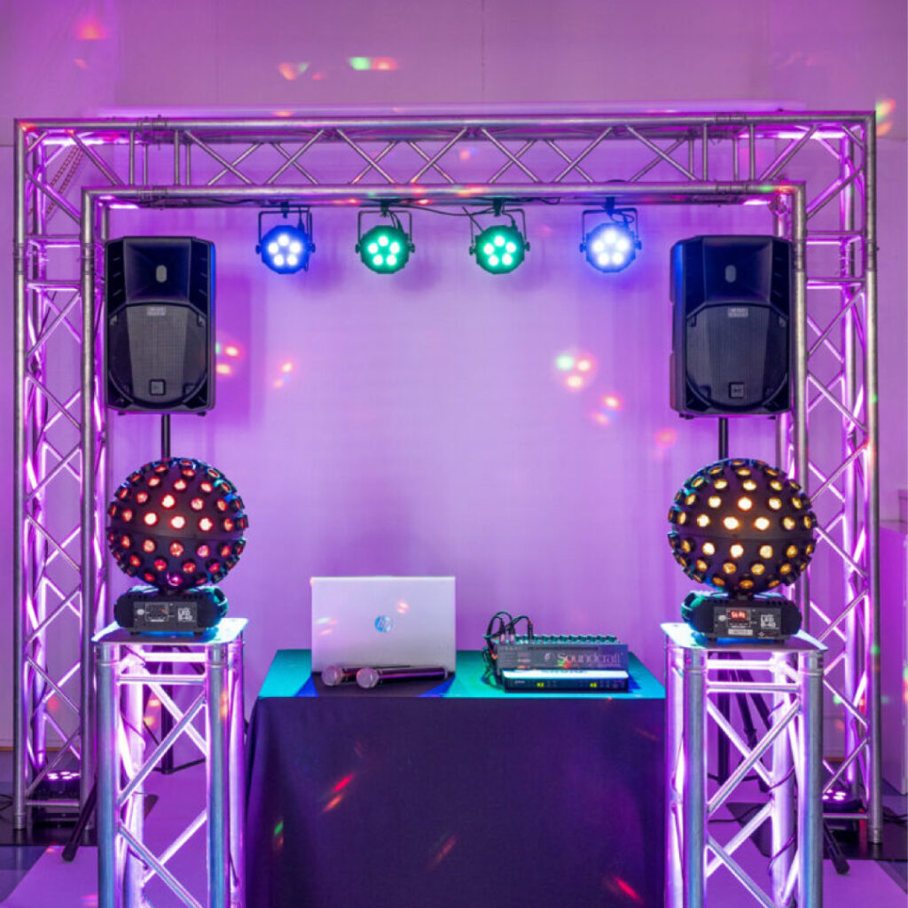 Valaistu trussikaari ja kaksi trussipylvästä, joiden välissä DJ-laitteet, kaksi suurta kaiutinta, kaksi diskopallovaloa, mikseripöytä ja kannettava tietokone. Yläpuolella neljä värikästä spottivaloa, taustalla violetti tunnelmavalaistus – DJ ja karaoke Turku sekä Varsinais-Suomen juhliin ja häihin.